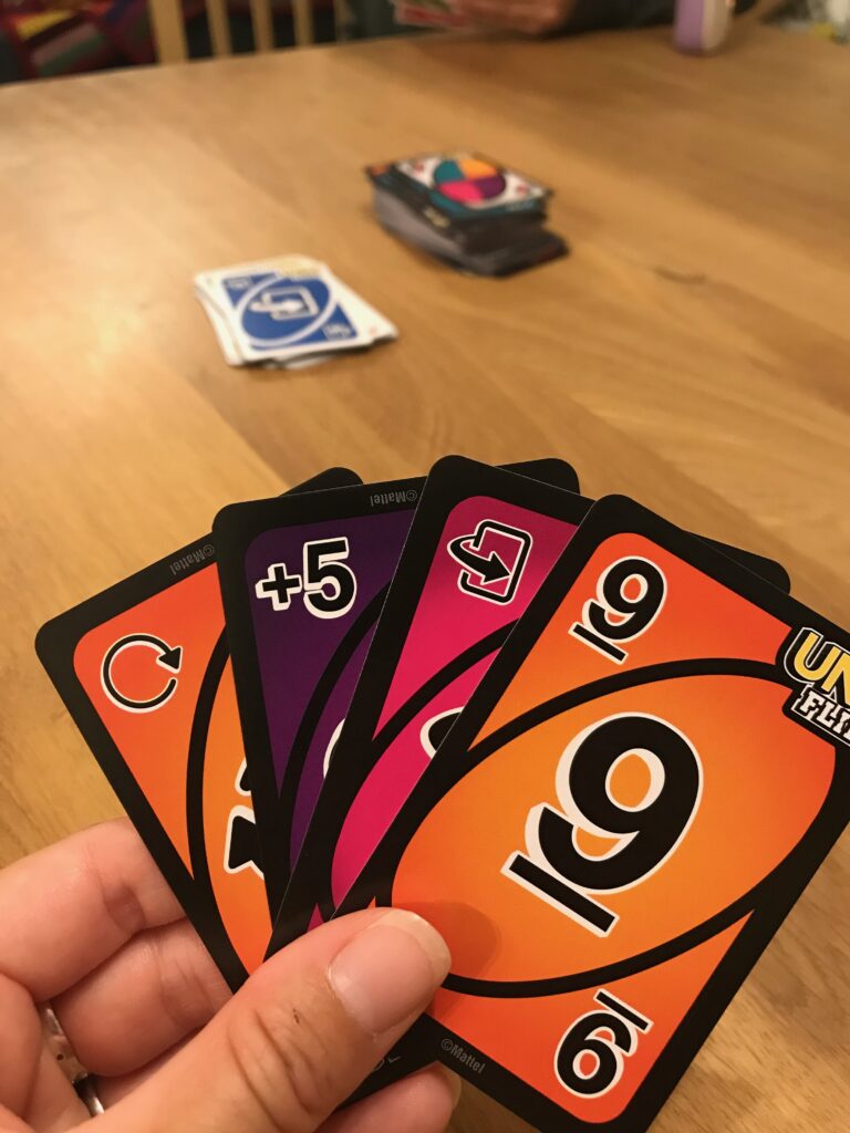 In the foreground of teh picture is a hand of Uno cards showing teh dark side of teh cards in the game Uno Flip. In the background you can see a draw and discard pile on the table.