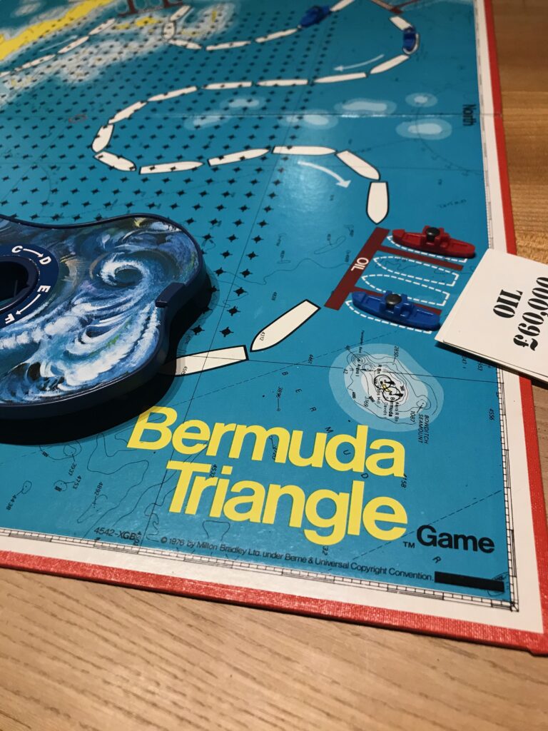 A corner of the board for Bermuda Triangle where it shows the name of the game. a couple of ships in port, some cards and the cloud covering part of the route.