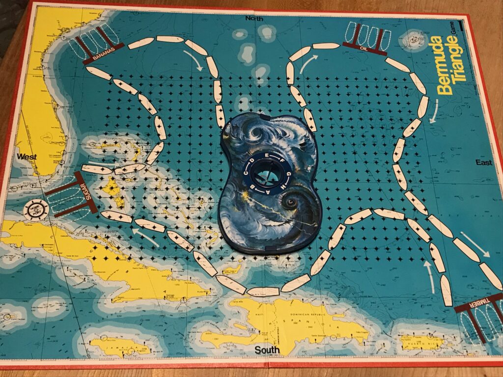 The board for Bermuda Triangle. Most of the board is sea with a track of boat shaped "squares" going round in a route, touching 4 ports on land. In the middle of the board is a grid and on this sits a strange plastic cloud shape which is the Bermuda Triangle in the game.