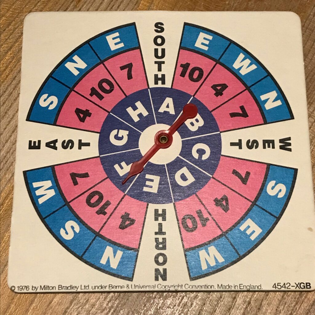 A cardboard spinner showing three rings. The inner one with a letter A-G. The second with a number: 4, 7 or 10 and then an outer one showing N, S, E or W. At the main compass points the second and third rings are merged and just say North, South, East or West.