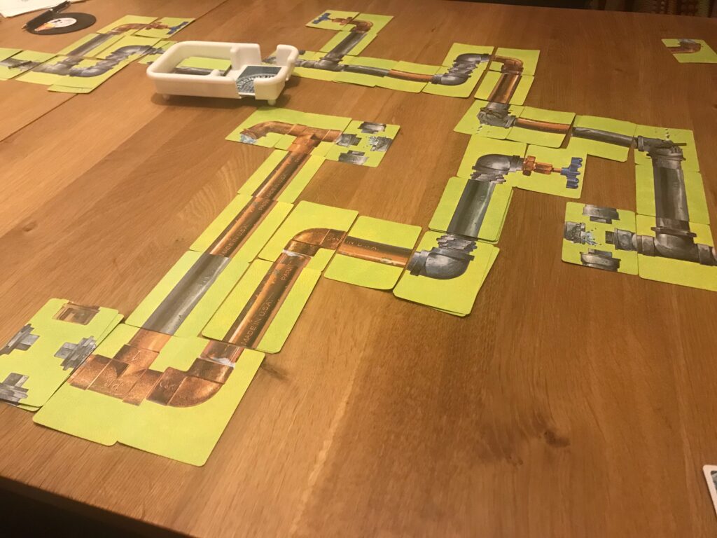 A section of pipework laid out in card form in the game waterworks. Also seen in teh picture is the plastic bathtub for drawing and discarding cards.