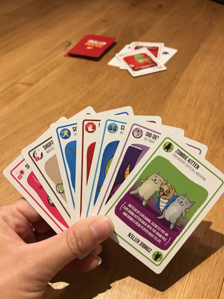 A hand of cards in Beth game Zombie Kittens. In teh hand you can see an array of different cards with a Zombie Kitten card at the front. In the background on the tabletop is a draw and discard pile.