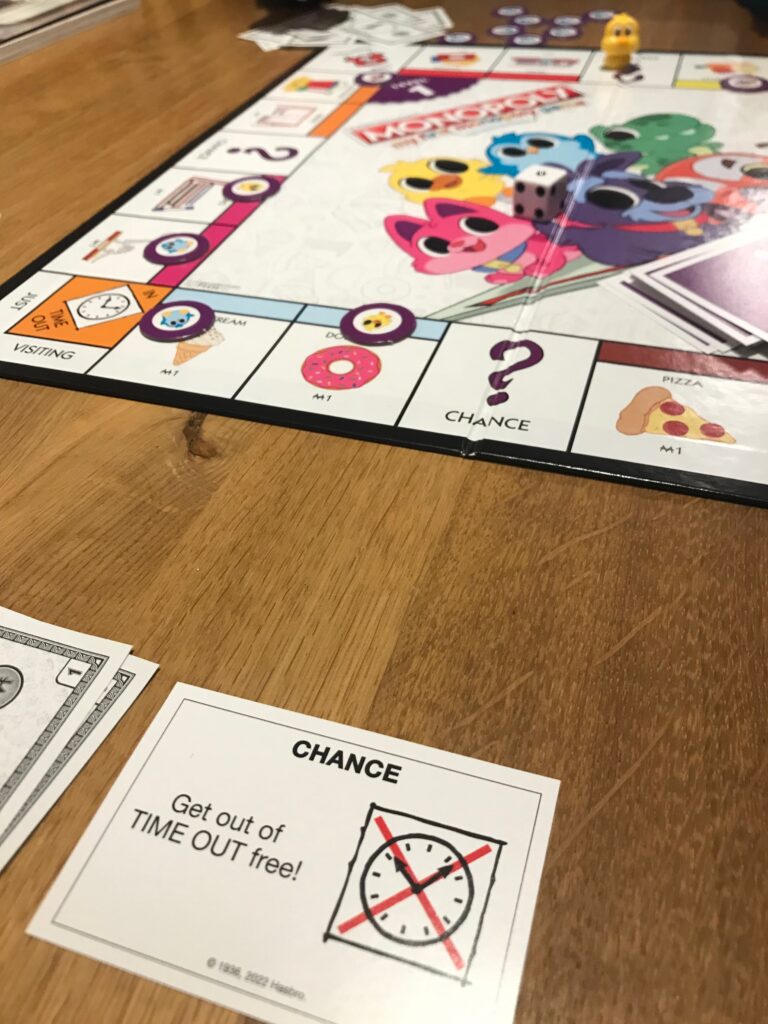 The My First Monopoly board is in the distance with a game in progress. In the foreground you can see a Get out of Time Out Free chance card and the corner of some Monopoly money. On the board you can see several of the Level 1 sold signs on properties.