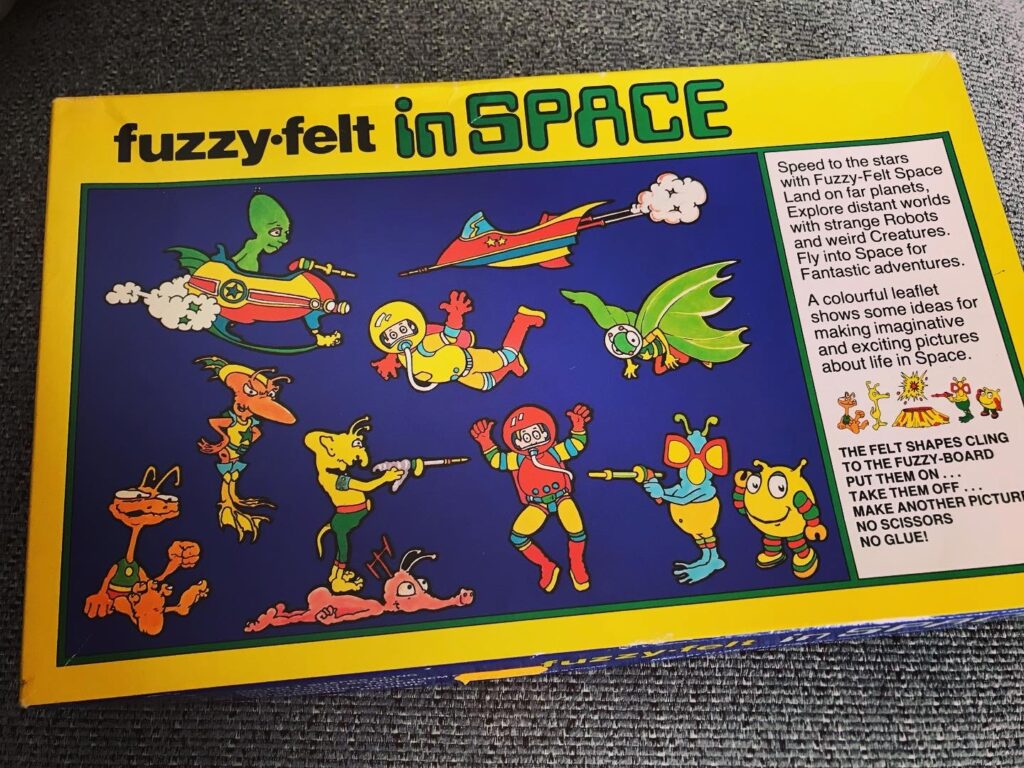 The box for Fuzzy Felt in Space. A yellow border and a dark blue background showing some of teh felt pieces in the box displayed in a space scene. 