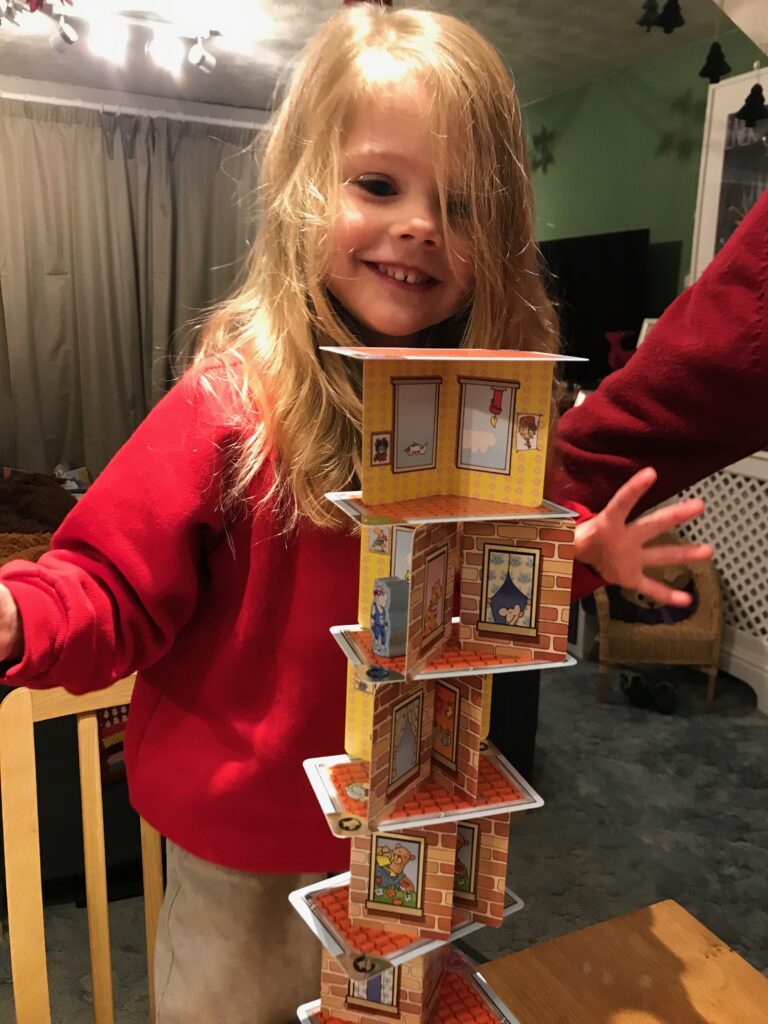 A 4 year old blonde girl is standing on a chair with a smile on her face and her hands outstretched as she's just placed a next level on Rhino Hero's tower.The tower that can be seen is at least six storeys high.