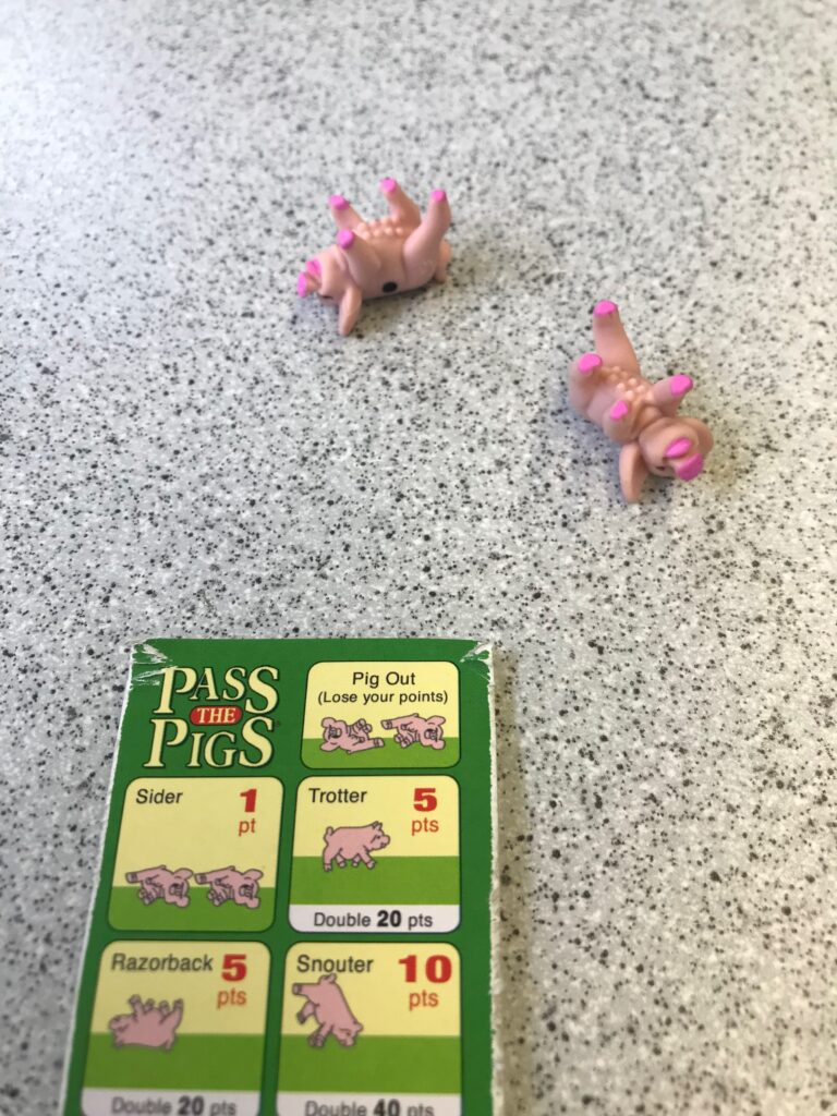 Two small pink pigs lying on their backs on a train table, with a Pass the Pigs quick reference scoring guide in shot showing that this is a double razorback and the player has scored 20 points.