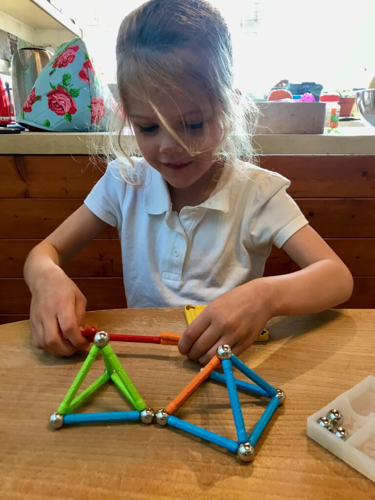 A blonde four year old girl smiling as she plays with Geomag. Infant of her on the wooden table are two pyramids built with Geomag. One with a square base and the other with a triangular base.