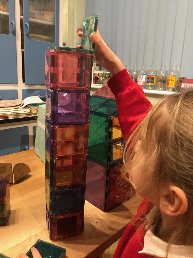 A blond five year old girl wearing a red jumper reaching up to place a tile at the top of a tower of 6 blocks, each made from magnetic tiles. 