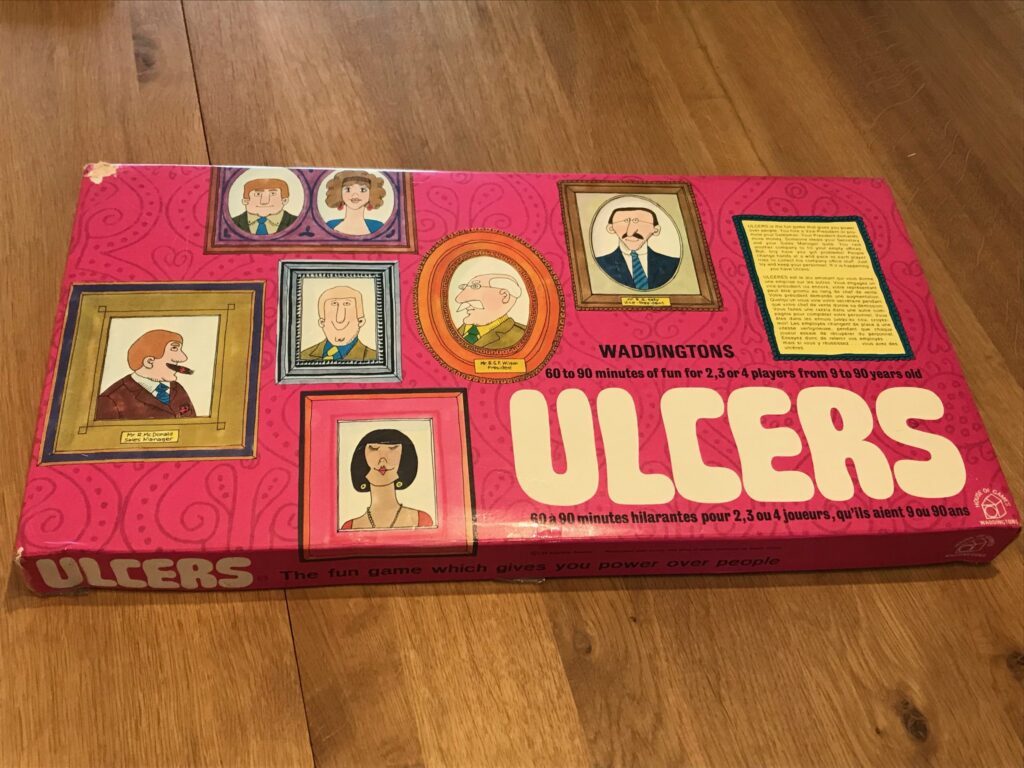 The box of Ulcers sitting on a wooden table top. The box is bright pink in colour with Ulcers written in bubble writing. Also visible are a series wow carton style pictures of personnel in ornate frames with their business positions written below them.