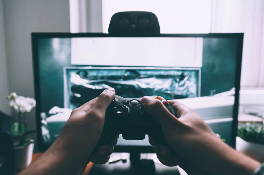 A muted tone photograph showing hands in the foreground holding a game controller whilst in the background is a large computer screen. The pose suggests the player is involved in a game but it is not possible to see clearly what they are playing. Illustrating an article entitled: Gift ideas to get boys away from screens this Christmas