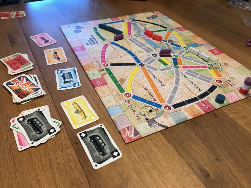 A game of Ticket to Ride London in action. On the board are several claimed routes and on the table around the board you can see an array of transportation tickets featuring a black cab, a yellow submarine, a blue milk float, an orange car and a pink racing car.